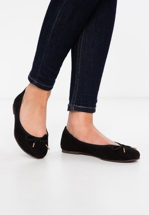 Gambe in jeans skinny blu scuro con ballerine in camoscio nero con piccoli fiocchi decorativi su sfondo di pavimento bianco.