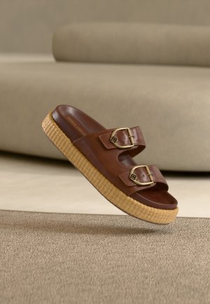 Καφέ δερμάτινο πέδιλο τύπου slide με δύο χρυσές αγκράφες και χοντρή πλεκτή σόλα πλατφόρμας επάνω σε μπεζ χαλί και φόντο καναπέ.