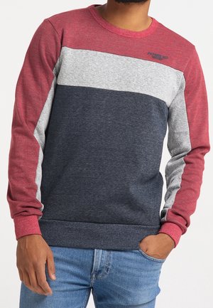 Mann trägt einen langärmeligen Farbblock-Sweatshirt in Rot, Grau und Navy mit einer Hand in der Tasche, kombiniert mit blauen Jeans.
