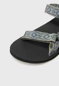 Flate, svarte sandaler med to mønstrede stoffstropper i blått og beige, med en teksturert såle for stabilitet.