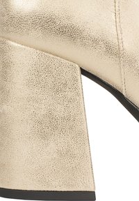 Scarpa con tacco alto color oro con superficie testurizzata, caratterizzata da un tacco a blocco affusolato e suola nera. Inquadratura ravvicinata del dettaglio del tacco.