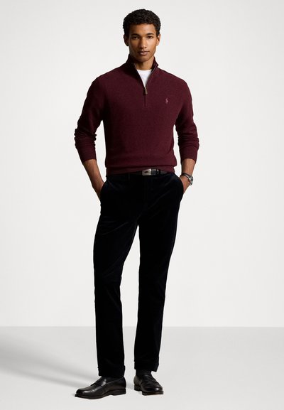 Pull zip-up bordeaux en tissu doux, associé à un pantalon en velours noir et des mocassins noirs. Coupe standard, avec un logo discret sur la poitrine.