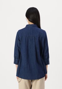 Dark blue denim shirt with a collar, buttoned cuffs, and a back pleat. The hem is slightly curved for a relaxed fit.