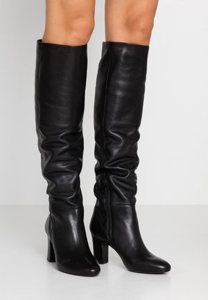Bottes en cuir noires à talons hauts, longueur genou, avec un design légèrement décontracté et une fermeture éclair latérale, portées sur des jambes nues.