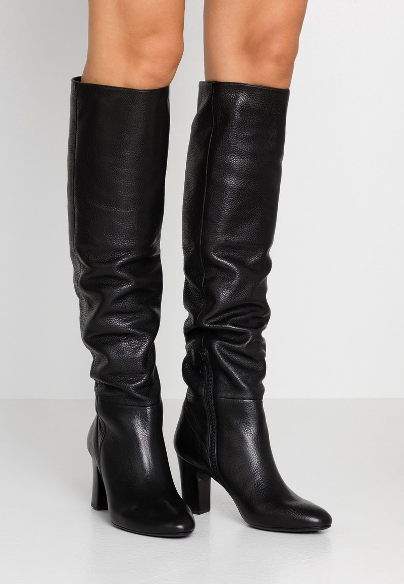 Bottes en cuir noires à talons hauts, longueur genou, avec un design légèrement décontracté et une fermeture éclair latérale, portées sur des jambes nues.