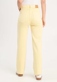 Jean jaune taille haute à jambes larges, avec poches arrière et une plaque en cuir beige au niveau de la taille, fabriqué en denim.
