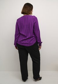 Blusa a righe viola con maniche lunghe, caratterizzata da un orlo arrotondato, abbinata a pantaloni neri e scarpe con tacco nere. Tessuto di trama liscia.