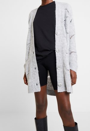 Osoba nosící dlouhý šedý pletený kardigan s velkými knoflíky přes černé tričko, černé krátké kalhoty a vysoké černé boty.