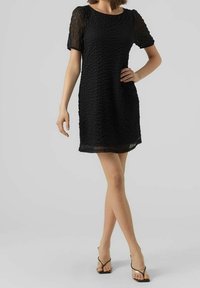 Robe noire courte en tissu texturé, à manches courtes et décolleté rond. Portée avec des sandales noires à brides, le modèle pose debout.