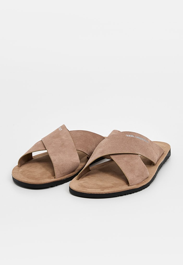 Sandalen aus beigem Wildleder mit einem über kreuz verarbeiteten Riemendesign, flacher schwarzer Sohle und weichem, strukturiertem Fußbett. Dezente Markierung oberhalb des Riemens.