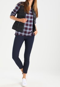 Czarna bezrękawnikowa kamizelka puchowa na granatowej i czerwonej koszuli w kratkę, w połączeniu z granatowymi dopasowanymi spodniami i białymi trampkami. Gładkie tekstury i swobodny styl.