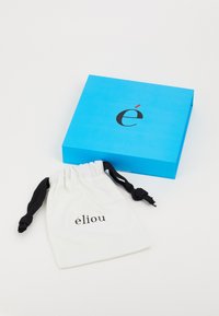 Boîte bleue avec le texte noir "é" et un accent rouge ; à côté, un sac à cordon blanc avec "élio" imprimé en noir.