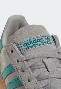 Grijze suède sneaker met teal strepen, tekst "adidas HANDBALL SPEZIAL" op de tong en een geprofileerde rubberen zool. Veters zijn lichtgrijs.