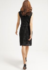 Černé přiléhavé šaty s ribbovaným vrškem, květinově texturovanou spodní částí, po kolena a se zadním rozparkem, doplněné černými vysokými podpatky.