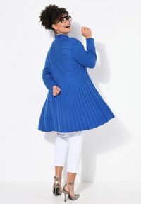 Largo cárdigan azul con pliegues y un diseño texturizado, combinado con pantalones blancos recortados y zapatos de tacón metálicos.