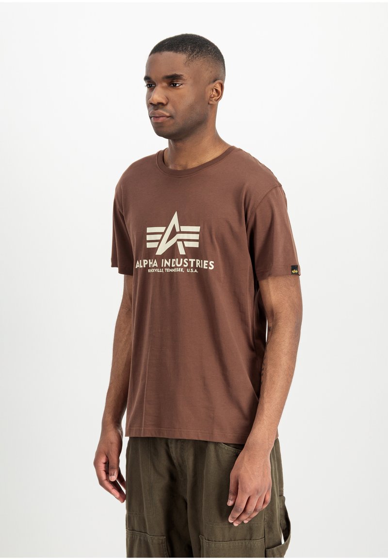Jeune homme noir portant un t-shirt marron Alpha Industries et un pantalon cargo vert olive, debout devant un fond blanc uni.