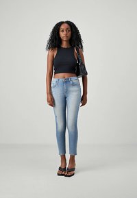 Jonge vrouw met krullend haar, gekleed in een zwart mouwloos crop top, lichtblauwe skinny jeans, zwarte sandalen en een zwarte schoudertas.