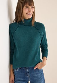 Maglione a collo alto color teal con texture a coste, caratterizzato da maniche raglan e una vestibilità ampia, abbinato a jeans blu. Aspetto liscio e accogliente.