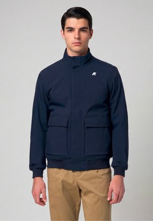 Jonge man met donker haar draagt een marineblauwe jas met hoge kraag en grote voorzakken en beige broek, staand tegen een lichte achtergrond.