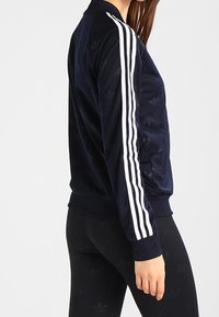 Marinblå bomberjacka med en slät tygstruktur, med vita ränder längs ärmarna och subtila mönsterdetaljer. Accenterad med en ribbad krage.