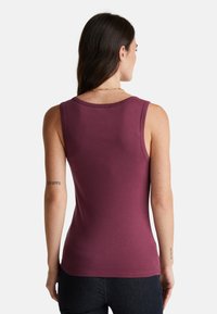 Bordeaux tanktop met een ronde halslijn, mouwloos ontwerp en een gladde textuur. Opvallende stiksels langs de randen. Draagt met een donkere spijkerbroek.