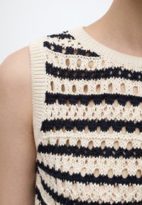 Αμάνικη πλεκτή μπλούζα με οριζόντιες ρίγες σε χρώματα κρεμ και μαύρο, με διάτρητα σχέδια, φορεμένη από άτομο με ανοιχτόχρωμο δέρμα.