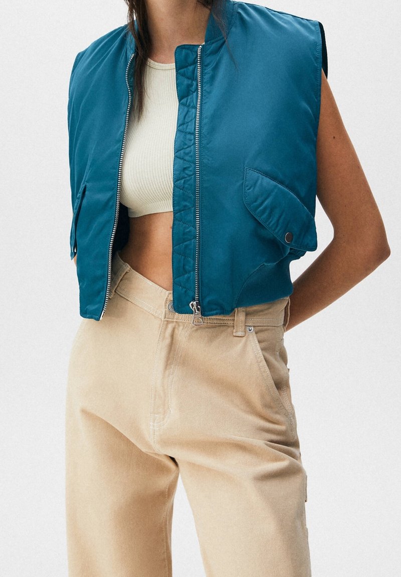Femme portant un gilet sans manches turquoise zippé, un crop top blanc et un pantalon beige taille haute, les mains dans le dos sur fond blanc.