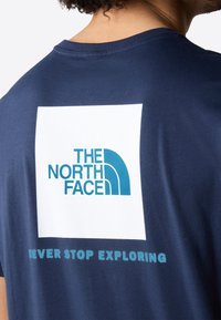 Granatowy bawełniany t-shirt z białym kwadratem i niebieskim logo z napisem "THE NORTH FACE" oraz tekstem "NEVER STOP EXPLORING" poniżej.