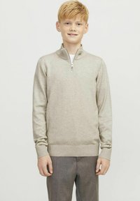 Svetlo bež zip-up jopič iz mehkega pletenega materiala, z ovratnikom in rebrastimi manšeti ter robom, v kombinaciji s sivoh hlačami.