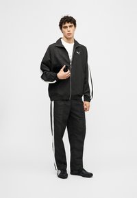 Νέος άντρας που φοράει μαύρη φόρμα Puma με λευκές ρίγες, κρατώντας ένα smartphone, σε απλό φωτεινό φόντο.