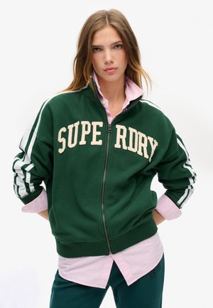 Sweatshirt zippé vert avec "SUPERDRY" en lettres crème. Rayures blanches sur les manches et poignets côtelés. Porté par-dessus une chemise rose clair.