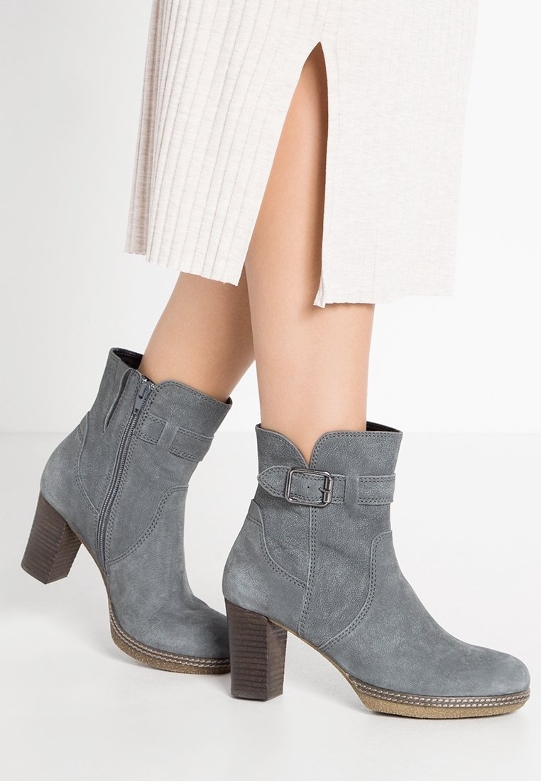 Femme portant des bottines en daim gris clair avec talons blocs et boucles latérales, associées à une jupe de couleur claire avec une fente avant.