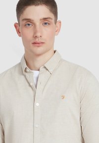 Chemise beige à boutons fabriquée en tissu texturé, avec un t-shirt blanc en dessous, un petit logo orange sur la poitrine gauche et un col classique.