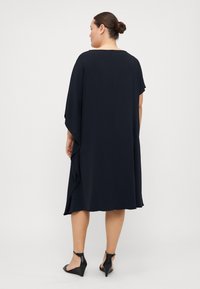 Marinekjole med løst snitt, korte ærmer og draperte sidesider. Glatt tekstur med rund halsudskæring. Bæres med sorte wedges.