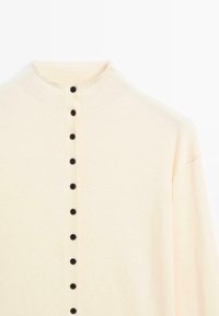 Pull beige clair avec un col montant, comportant une rangée de boutons noirs sur le devant. Texture douce, manches larges et design simple.
