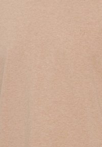 Tricou cu mâneci scurte, de culoare bej deschis, fabricat dintr-un material moale, cu un mic logo reliefat în zona stângă a pieptului.