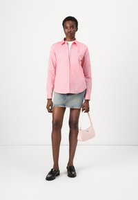 Camicia rosa chiaro con maniche lunghe, abbinata a una mini gonna di jeans. La modella tiene in mano una piccola borsa rosa e indossa mocassini neri.