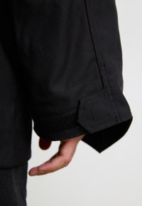 Mâneca unei jachete negre, cu un material texturat, închidere cu velcro și detalii de cusătură unghiulară la manșetă. O mână este parțial vizibilă.