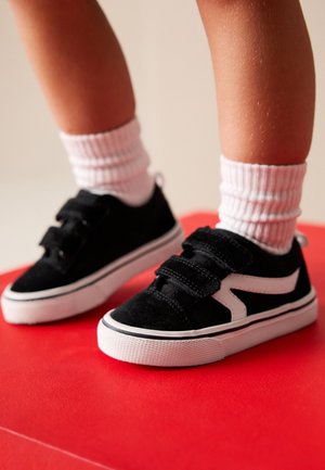 Enfant portant des baskets noires et blanches à scratch et des chaussettes blanches côtelées, debout sur une surface rouge.