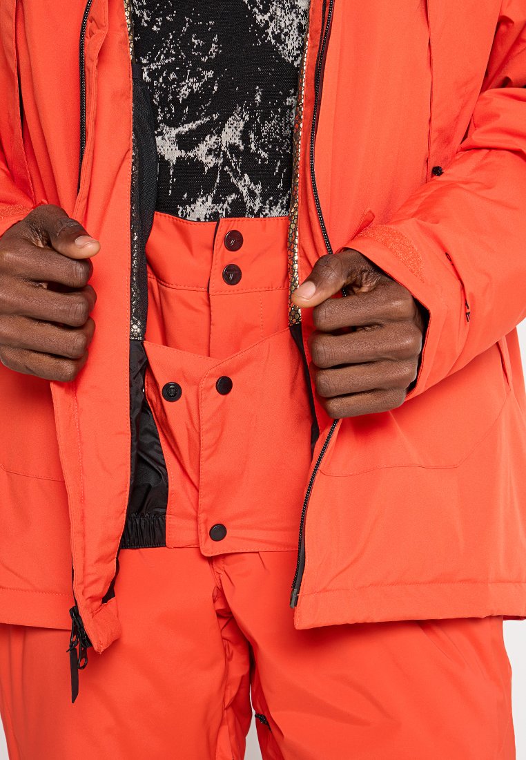 Helle orange isolierte Jacke mit einer schwarzen, zu öffnenden Innenschicht und passenden orangenen Hosen, die über Druckknöpfe und einen elastischen Bund für optimale Anpassung verfügen.