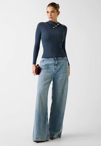 Marineblauwe geribbelde gebreide top met een diagonale kraag en gouden accenten. Lichtblauwe jeans met wijde pijpen en een ontspannen pasvorm. Wijnrode clutch in de hand.