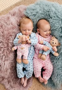 Two infants on fluffy rugs, one in a blue star-patterned onesie holding a teddy, and another in a pink star-patterned onesie with a teddy.