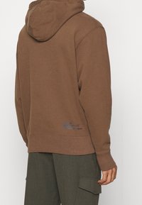 Sudadera con capucha marrón con un acabado texturizado, que presenta puños y dobladillo de canalé. Incluye un pequeño detalle del logo azul en la parte posterior.