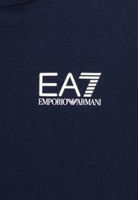 Marinblått tyg med vit "EA7 Emporio Armani" logotyp som har en stiliserad siffra 7 med horisontella ränder.