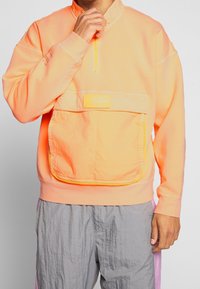 Pullover arancione brillante con colletto alto, zip a quarto e grande tasca frontale. Tessuto texture con accenti gialli a contrasto e dettagli cuciti.