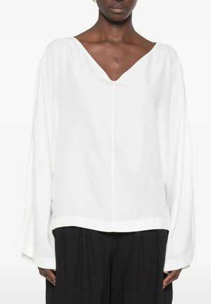 Persona che indossa una blusa bianca larga a maniche lunghe con scollo a V e pantaloni neri a gamba larga, mostrata dal collo ai fianchi su sfondo bianco.