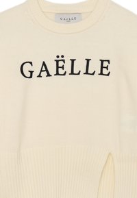 Maglione a maglia color crema con accenti a costine. Presenta la parola "GAËLLE" ricamata in nero sul davanti. Collo rotondo classico.