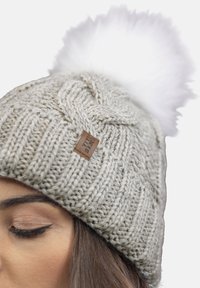 Berretto lavorato a maglia grigio chiaro con un motivo testurizzato, dotato di un morbido pom-pom bianco sulla sommità e di un'etichetta in pelle marrone.