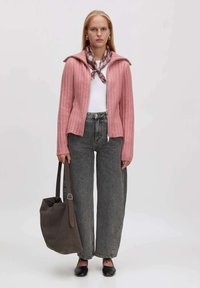 Frau trägt eine rosa Reißverschluss-Cardigan, ein weißes Oberteil, graue weit geschnittene Jeans, einen karierten Halstuch, schwarze Ballerinas und hält eine große braune Tasche.