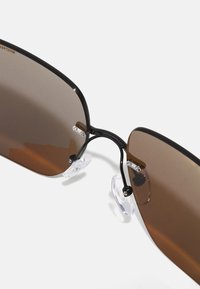 Ochelari de soare cu tentă maro, cu ramă din metal negru și suporturi pentru nas transparente. Lentile dreptunghiulare mai mari, cu hardware minim și un design elegant.